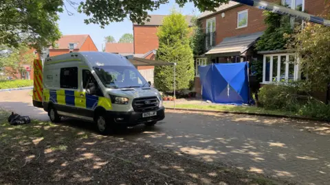 Laura Foster/BBC A police car, outside a house, parked on a road, with a blue police tent outside the house. There is also police tape near the top of the image. The house is mostly brick, with white window frames and foliage. Other houses are to the left. 
