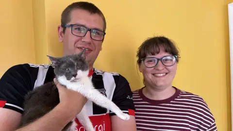 BBC/Lucy Ashton A man with close cropped brown hair has glasses and a black, white and red football shirt. He is holding a white and grey cat. Next to him is a woman with a brown fringe, glasses and maroon and white striped top. They are both smiling and stood in front of a yellow wall
