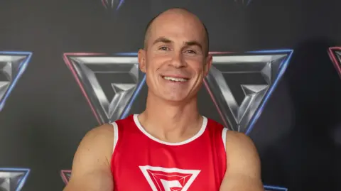 Mark Billings is smiling at the camera. He is wearing a red and white vest top with the Gladiators logo on the chest. He has his arms crossed.