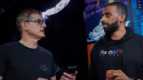 Netflix Myron Gaines, wearing a Fresh & Fit podcast hoodie, listens as Louis Theroux interviews him inside the influencer's home studio. He holds a can of energy drink in one hand, Theroux holds a phone, ready to play a clip.