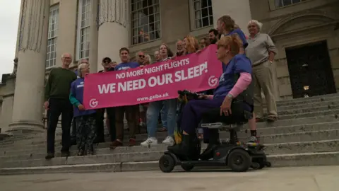 A group of 20 protesters from GALBA gather on the steps of Leeds Civic Hall to protest against night flights at Leeds Bradford Airport. They're holding a big, pink banner with the words We Need Our Sleep, No More Night Flights