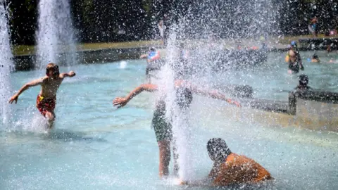 Teenage boys playing in a large fountain of a splash park. Their faces are obscured from the camera. One boy has his face in a large water fountain spray. It is a bright sunny day.