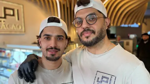 Two men stand next to each other wearing white t-shirts with a logo on. The are wearing white caps which are back to front and the taller man on the left has his arm around the smaller man's shoulder.