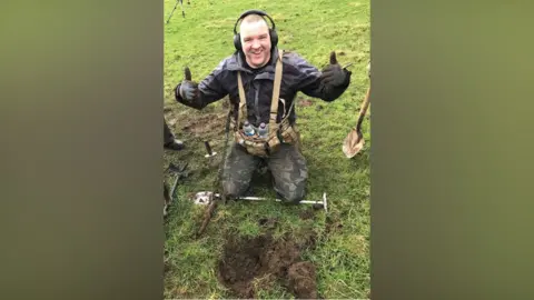 Chris Linton is kneeling on the grass in front of a hole in the ground. He is surrounded by metal detectoring equipment and tools to dig. He is wearing headphones, a black and grey jacket, camouflage trousers and a harness with water bottles. He is looking at the camera and smiling with two thumbs up