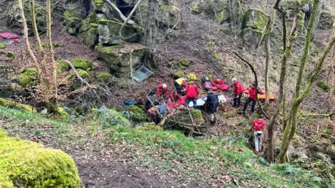 Rescue teams are pictured in a dense woodland. They are wearing red jackets and are surrounding a casualty while they treat them