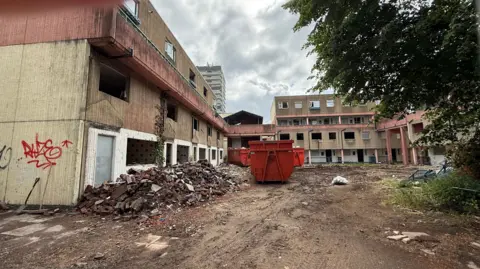 A demolition site, showing lots of derelict deteriorating buildings with a red skip infront of it 