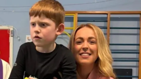 A woman with long blonde hair wears a pink top as her small ginger haired son sits on her lap. She is looking at the camera while he looks of to the left. He wears a black long-sleeve t-shirt. Behind them is a blue wall with wooden beams on the wall. 