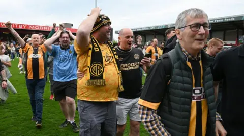 PA Media Fans in amber and black United colours seen walking on the pitch with happy, emotional expressions.