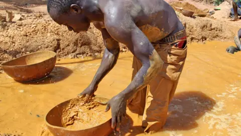 AFP via Getty Images A bare-chested man stands ankle-deep in yellow-coloured water, holding a bowl with some mud in it, panning for gold.