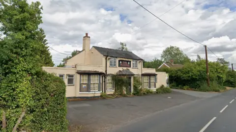 A cream coloured pub over two floors with boarded up windows and a black sign that reads: "The Plough." 