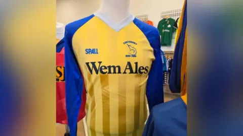 12th Man Retro A yellow and blue shirt sits on a mannequin. It says Wem Ales and has a logo and date on the right breast. Other different coloured shirts can be seen behind it.