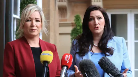 PA Michelle O'Neill and Emma Little-Pengelly speaking at a row of media mircophones
Michelle is in a red suit blazer and emma little pengelly is in a blue blazer and top 