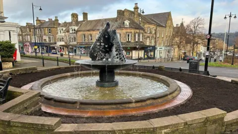 A fountain made from steel in the shape of two leaves, with water flowing through it into a stone circular pool within a flower bed. It stands at the top of a street with Victorian stone shops in the background. 