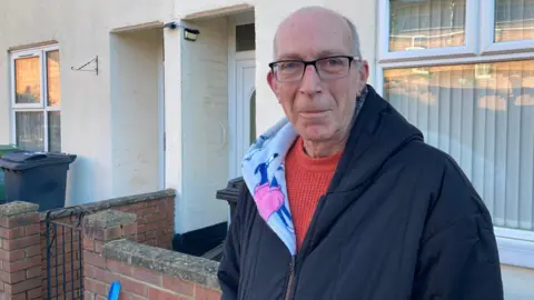 An older man with short grey hair at the back of his scalp, black rectangular rimmed glasses, a red jumper and a black hoodie with a fleecy, patterned interior stands outside of his terraced home in a city suburb on a sunny winter morning,