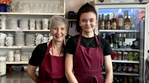 Ellis Karan/LDRS Two women wearing maroon aprons standing behind a cafe counter, smiling