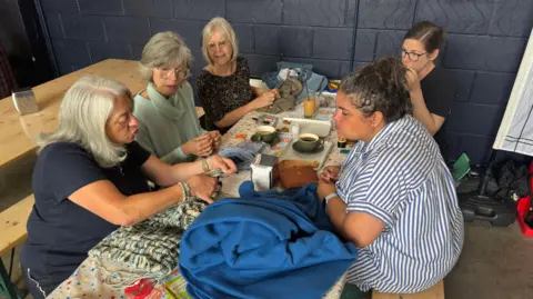 Five women sit around a long table. They are drinking coffee, chatting and doing various crafts, including sewing and knitting. One women is demonstrating a technique to others in the group.