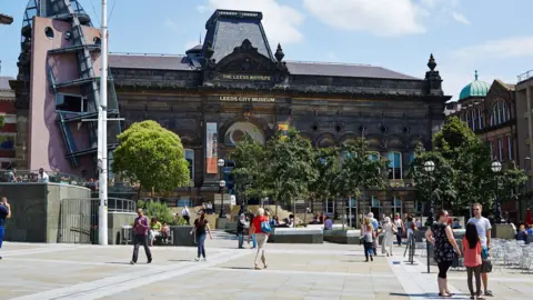 The front of Leeds City Museum. It is a sunny day, with people pictured walking in front of the building.