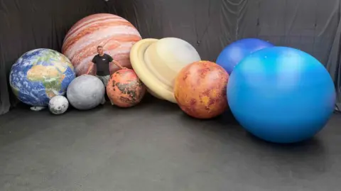 Glow Inflatables A man stands in a large room filled with inflatable planets. The planets are from our solar system and are different colours and sizes.