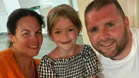 Family photo Emma (left) has dark hair, pulled back into a ponytail, and is wearing a bright orange top and smiling at the camera. She is next to Ruby (centre), who has her hair up and is smiling at the camera. Ruby is wearing a black and white gingham outfit and is next to her dad Dean (right), who is also smiling and wearing a white T-shirt.