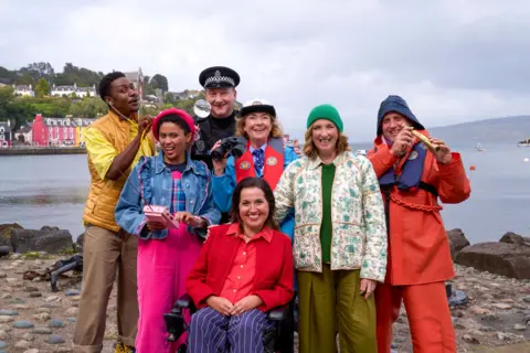 Lion TV Media Scotland/BBC The cast of the Balamory revival stand on the harbour dressed as their characters - Ollie (CARL SPENCER), Ava Potts (DANIELLE JAM), PC Plum (ANDREW AGNEW), Edie McCredie (JULIET CADZOW), Penny Pocket (KIM TSERKEZIE), Miss Hoolie (JULIE WILSON NIMMO), Harbourmaster (WILLIAM ANDREWS)
