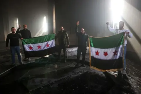 Five male Syrian rebels hold two Syrian flags with their arms raised in celebration standing over the burned gravesite of Hafez al-Assad.