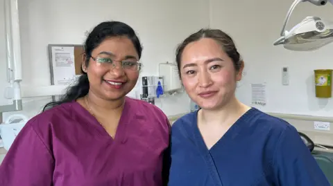 Ellen Knight/BBC Raj and Dr Gurung stood side by side. Raj is wearing a berry-coloured scrub top, and Dr Gurung is wearing dark blue scrubs. Raj has dark wavy hair, tied back in a half-up hairstyle. Dr Gurung has her dark hair tied back into a bun. They're both stood in a treatment room, in front of the dentists chair. The room has white walls and white ceilings. 