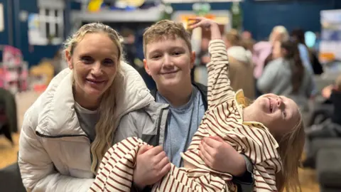 A woman with blonde hair wearing a white coat, stands next to a young boy who is in a grey t-shirt and black jacket, he is holding a young girl in his arms who is wearing a cream and brown striped top and tracksuit bottoms,