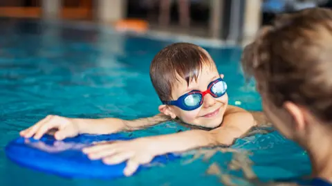 Getty Images Stock image of a young boy in a swimming pool. He has wet brown hair and is wearing blue goggles while holding a blue float. He is being instructed by a woman who is viewed from behind.