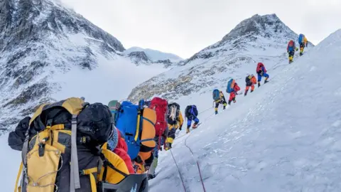 A line of mountain climbers hike across a snow-covered slope with mountain peaks in the background