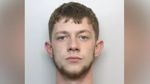 South Yorkshire Police Strachan stands against a plain, light‑coloured background. He has short, light brown hair that is neatly trimmed. His ears are visible, and he is wearing a simple light grey crew‑neck shirt.