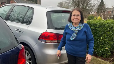 Hannah Gray/BBC Marbelia Davison pictured outside her home, beside her silver car. Marbelia is wearing a blue jumper, and patterned neckerchief. She has shoulder length hair and is wearing glasses. She is holding her car keys.