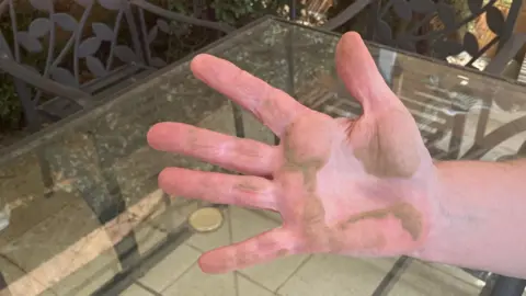 The hand of Sandra Caller has dust on it after she swipes her garden table with it. Her hand is raised to the camera, with a glass garden table in the background
