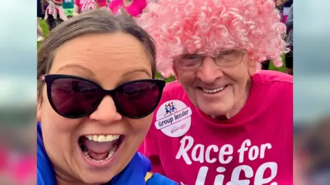 Julie Blackburn A smiling woman with tied-back brown hair and sun glasses smiles alongside an older man wearing a pink 'Race for Life' T shirt and pink curly wig. It is a selfie.