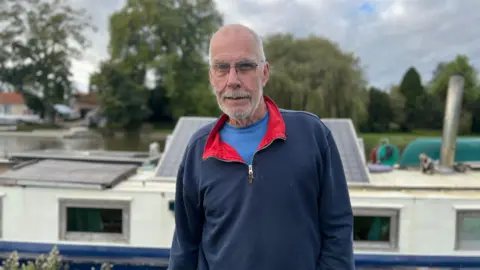 A grey-haired man wearing a blue sweatshirt looking straight at the camera. The roof of his boat and the river are in soft focus behind him.