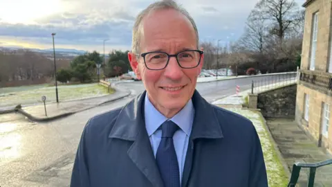 Prof Alan McNeill stands on the pavement with an old office building to his left and a grassy hill to his right and behind them. He is wearing a navy coat and a blue shirt and tie and glasses.