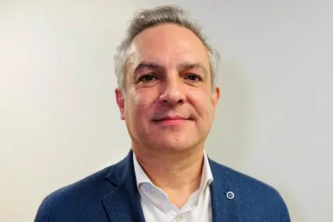 Chris Littlejohn, a man smiling a camera, wearing a blue suit jacket over a white short, unbuttoned at the colar.