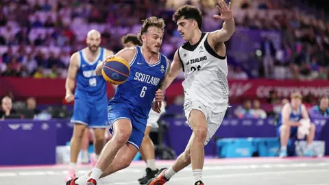 Getty Images Kyle Jimenez of Team Scotland - wearing an all blue basketball uniform - is challenged by Christian Martin of Team New Zealand, wearing all white. Other players can be seen in the background, while the crowd are blurry in the distance.