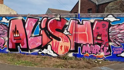 Family A large colourful graffiti-style mural displays the name Alisha Marie in pink and red lettering with decorative artwork and wing motifs on a wall beside a road.