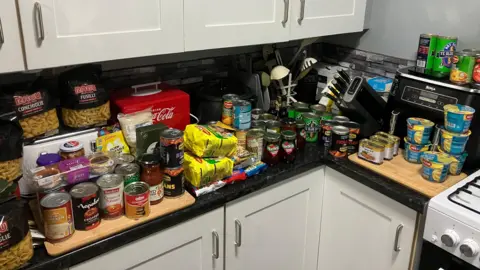 Stacks of food tins and packets on a black kitchen counter. They include pasta, fizzy drinks, beans and jam.