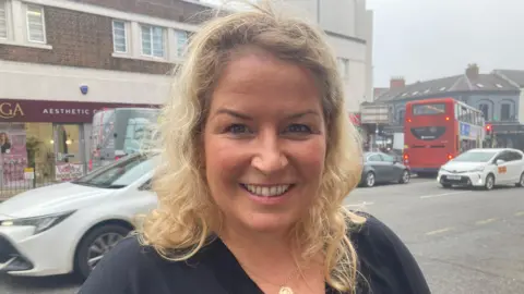 Tom Percival/ BBC A blonde haired woman in a black dress stands in front of a road with traffic driving past 