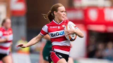 Mia Venner runs with a ball held to her body under her left arm, with her right arm held out during a match for Gloucester-Hartpury