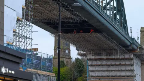 Kontraktor yang mengenakan pakaian hi-vis bekerja di perbaikan jembatan Tyne. Mereka berada di perancah yang didirikan di bagian bawah jembatan.