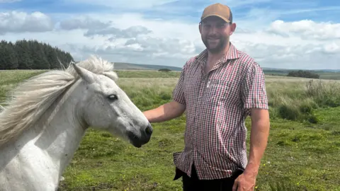 George Abel is wearing a red-and-white short-sleeved checked shirt and a tan baseball cap. He is standing in open moorland. His face is towards the camera but his body is pointing towards a grey pony which is standing in profile in the left of the frame. Abel's right hand is reaching out to pet the pony.