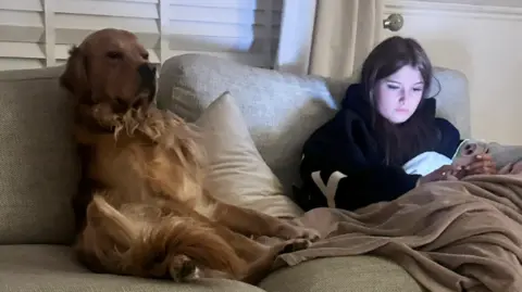 Girl Ellie sits on a sofa with golden retriever Louie next to her. She has brown hair and is wearing a dark hoodie with a brown blanket over her legs 