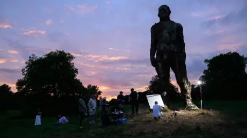 Nicholas Smith A film set in a field at dusk. There is a large sculpture of a man towering above the film set. People hold microphones and other filming equipment.
