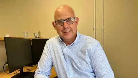 A man sits next to two black computer screens. He has short grey hair and wears black glasses. He wears a light blue patterned shirt and jeans. 
