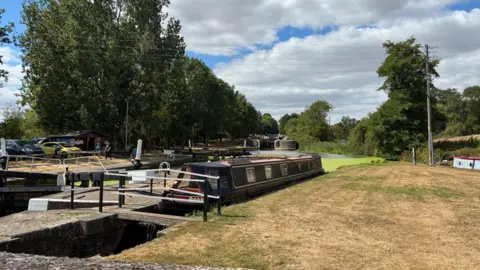 An image of Hatton locks with a canal boat parked next to one of the locks. dry grass on the tow path next to the canal. Much of the water is green with algae and the locks continue off into the distance with trees either side of the canal.