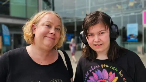 A closed mouth smiling Victoria Trattles who has blonde chin-length hair, wearing a black t-shirt with her daughter Maria who has dark shoulder-length hair and is wearing ear protectors and a black t-shite with a multi-coloured flower on it 