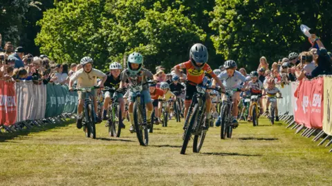Red On Sports/Angus MacKinnon A group of young bike riders speed across the grass with crowds lining the route beside them