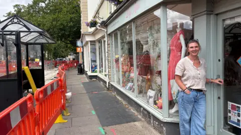 The store front of Lily's attic with the owner Tamsin stood out the front. Opposite the shop is a closed bus stop with orange fencing stretching down the street.
